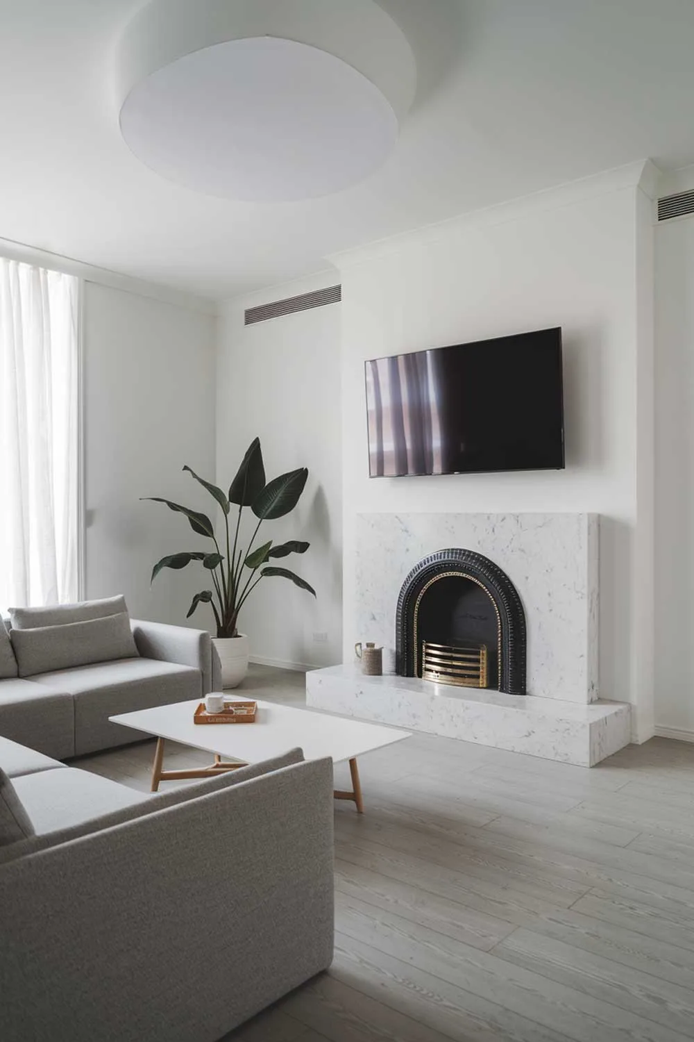 A minimalist living room with a white fireplace and a mounted TV. The room has a grey sofa and a white coffee table in front of the sofa. There is a potted plant near the fireplace. The walls and the ceiling are white. The floor is made of light grey wood.