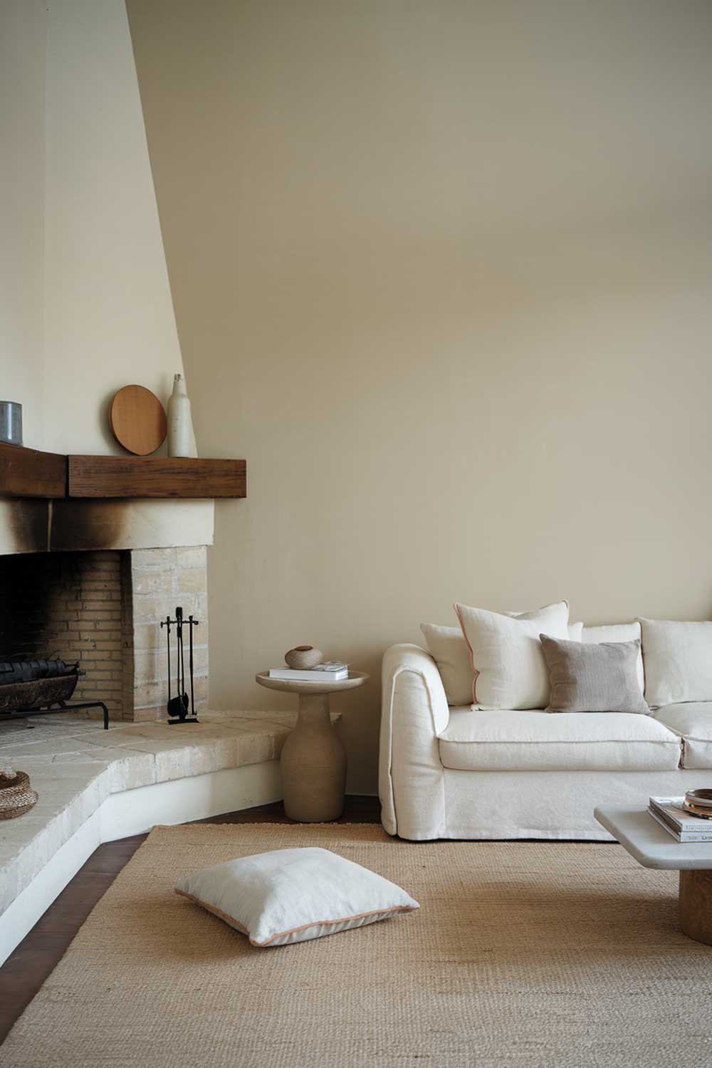 A minimalist living room with a corner fireplace. The room has a white sofa, a few scattered pillows, and a side table. The fireplace has a wooden mantel and a few decorative items. The floor is covered with a beige rug. The wall behind the sofa is painted beige.