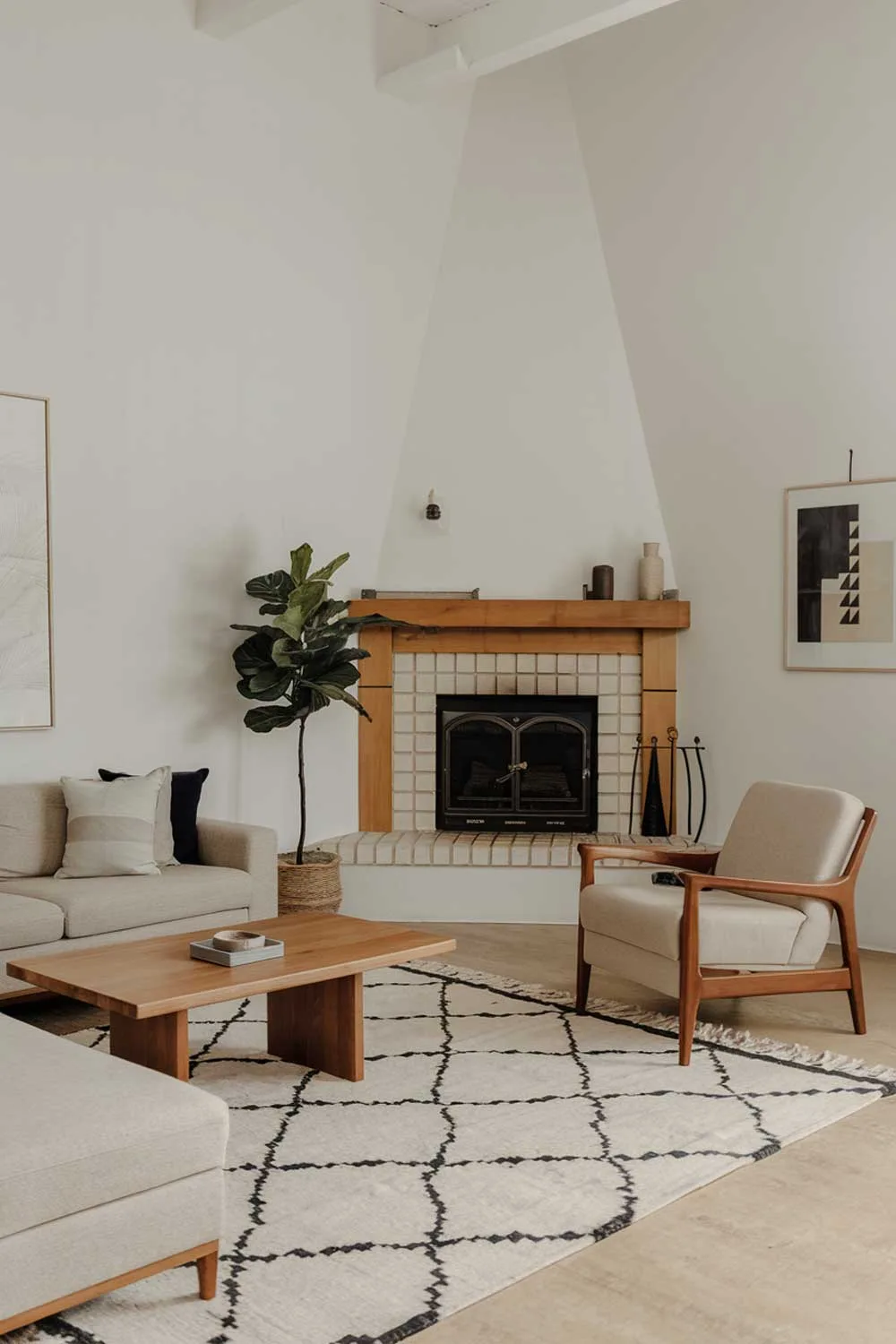 A minimalist living room with a corner fireplace. The room contains a beige sofa, a wooden coffee table, a beige armchair, and a potted plant. There is a rug with a geometric pattern in the middle of the room. The walls are white, and there are a few pieces of art hanging on them. The fireplace has a wooden mantel and a white insert.