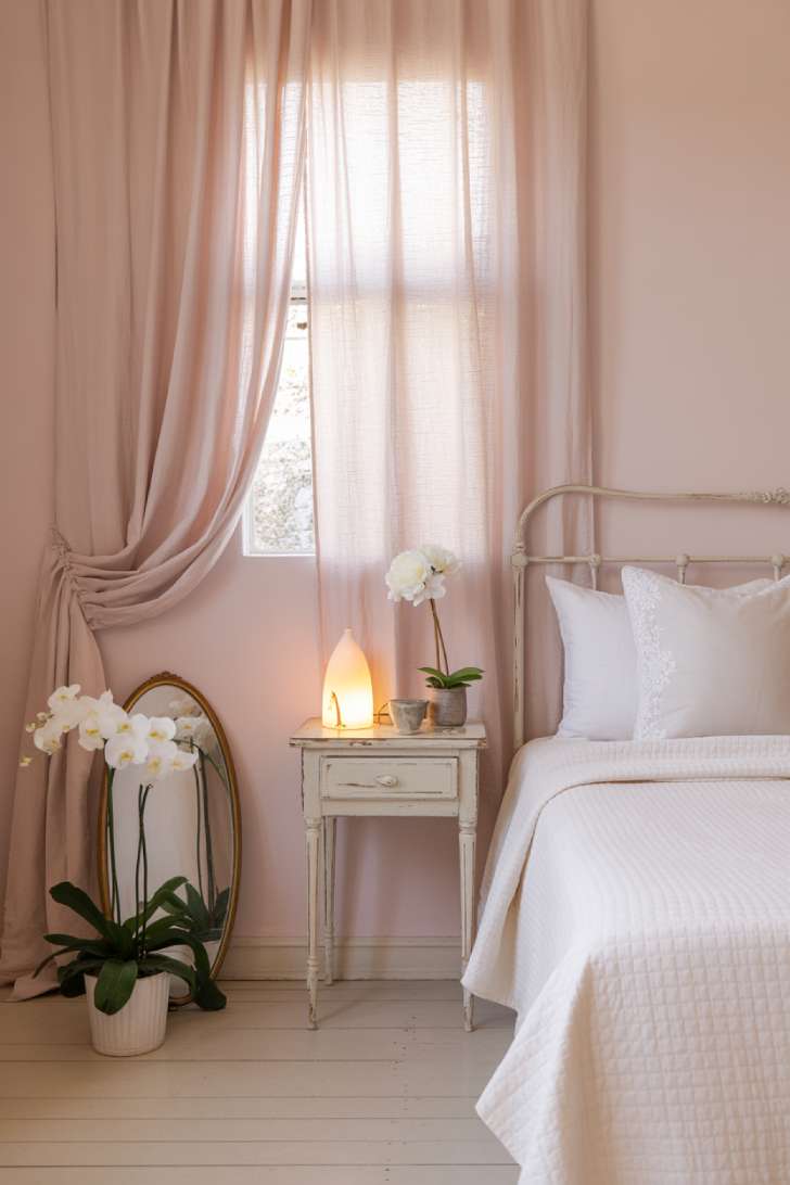 A romantic minimalist guest bedroom with an antique white iron bed, white broderie anglaise pillowcases, a blush-tinged wall, alabaster table lamp, and a fresh white peony in a bud vase