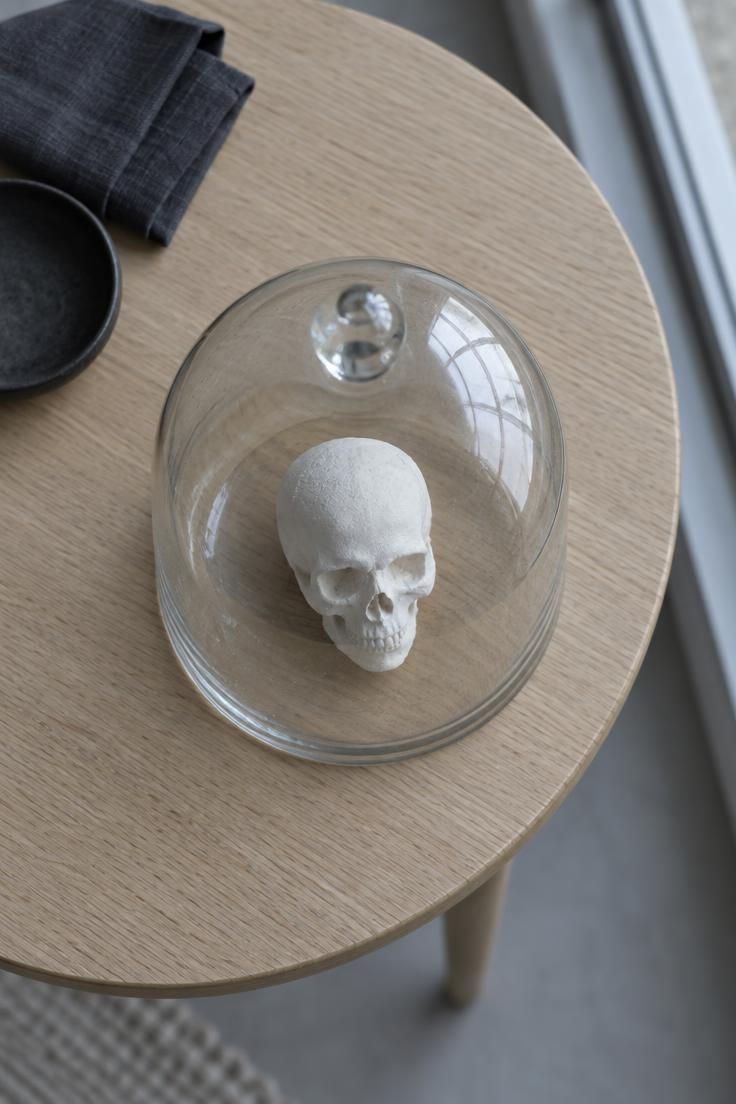 13 Warm Minimalist Halloween Ideas for a Calm Autumn Home 5 An overhead view of a glass cloche covering a small matte white skull form on a pale oak side table in cool daylight.