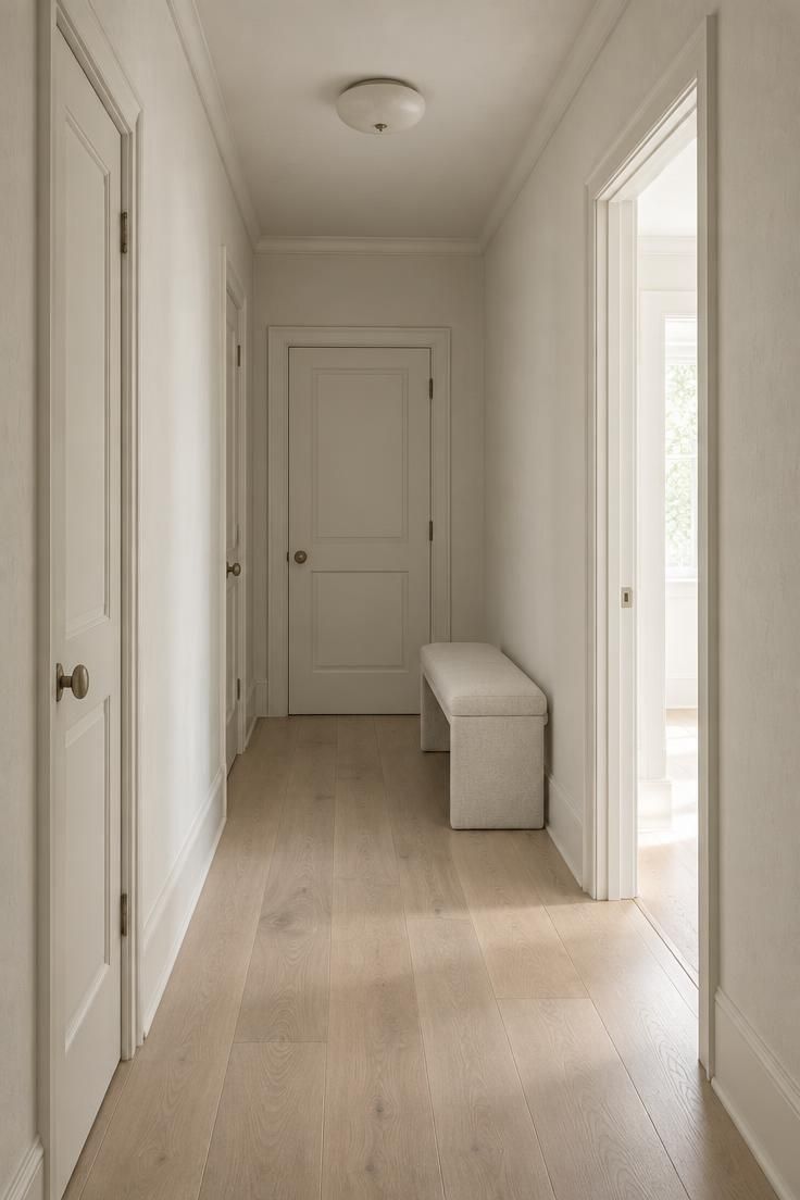 13 Warm Minimalist Hallway Ideas for a Calm Entryway 1 Narrow hallway with warm off-white walls, trim, doors, and ceiling painted one chalky tone, pale oak floor, and a single slim bench near the far end.