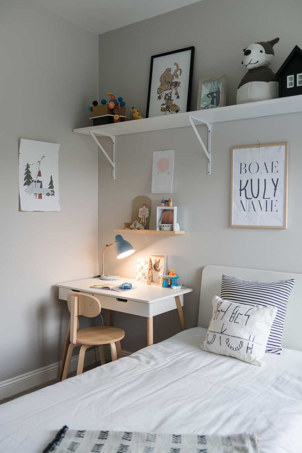 A cozy minimalist kids bedroom with a white bed, a few toys, and a shelf. There is a small desk with a lamp in the corner. The walls are painted light gray.