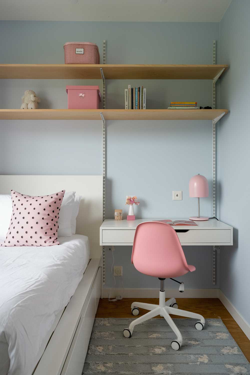 A minimalist girls bedroom with a white bed, a wooden shelf, a pink chair, and a small white desk. The room has a gray rug, a white lamp on the desk, and a pink lamp on the shelf. The walls are painted light blue.