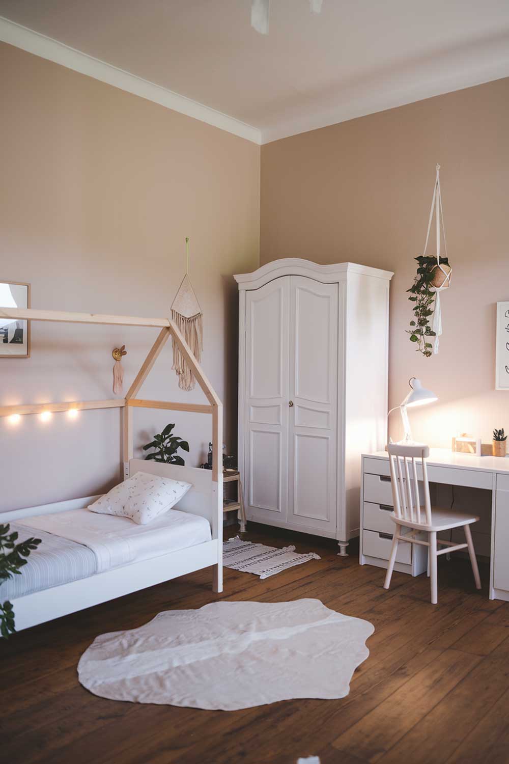 A minimalist girl's bedroom with a white bed, a white wardrobe, and a white desk. The bed has a wooden frame and is placed against the wall. There's a small plant near the bed. The wardrobe is placed near the desk. The desk has a chair and a lamp. The floor is wooden. The walls have a few items of decoration, including a framed picture, a string of lights, and a hanging plant. The room has a soft, warm lighting.