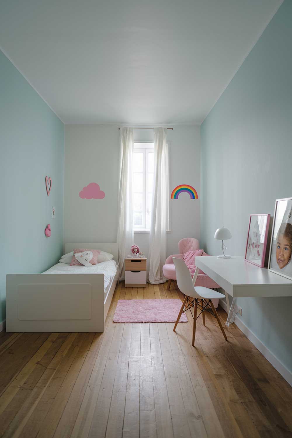 A minimalist girl's bedroom with a white bed, a small white desk, and a white chair. The room has a wooden floor and the walls are painted in a soft blue hue. There is a small pink rug near the bed. On the white wall opposite the bed, there's a pink cloud and a rainbow sticker. The room has a small window with white curtains. There's a lamp on the white desk. The room has a few personal items, such as a pink heart decoration and a framed image of a girl with curly hair.