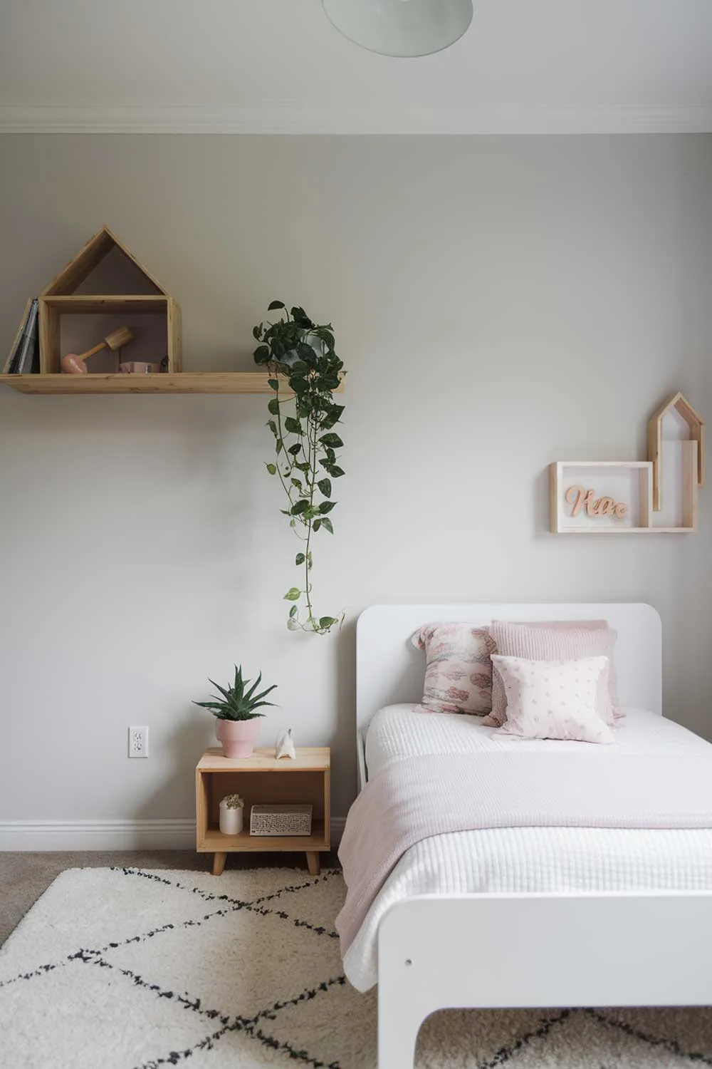 29 Minimalist Kids Bedroom Ideas: Simple Yet Playful Designs 14 A minimalist girls' bedroom with a white bed, a wooden shelf, and a small potted plant. The room has a light gray wall and a white ceiling. There is a white rug beneath the bed.