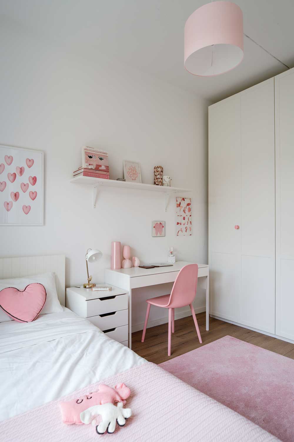 A minimalist girl's bedroom with a white bed, a few furniture pieces, and a few decorative items. The room has a few pieces of furniture, including a white bed with a pink heart pillow, a white wardrobe, a white desk with a pink chair, and a small white shelf. There are also a few decorative items, including a pink lamp, a pink rug, and a few pink and white decorative items on the shelf and desk. The walls are painted white.