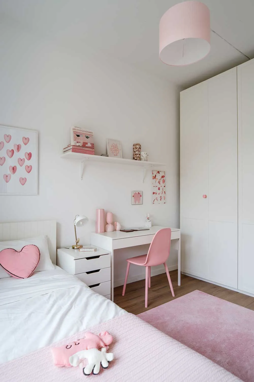 29 Minimalist Kids Bedroom Ideas: Simple Yet Playful Designs 15 A minimalist girl's bedroom with a white bed, a few furniture pieces, and a few decorative items. The room has a few pieces of furniture, including a white bed with a pink heart pillow, a white wardrobe, a white desk with a pink chair, and a small white shelf. There are also a few decorative items, including a pink lamp, a pink rug, and a few pink and white decorative items on the shelf and desk. The walls are painted white.