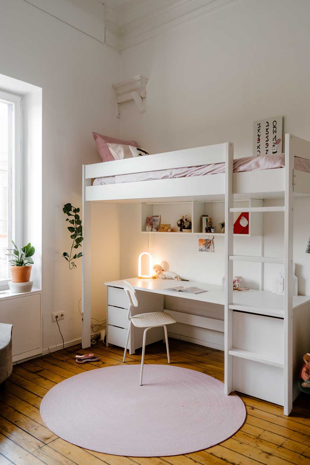 A minimalist girl's bedroom with a white loft bed, a small desk, and a chair. The room has white walls and a wooden floor. There is a small plant near the window. The room has a few personal items, such as a lamp and a doll.