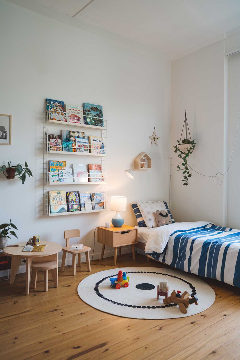 A minimalist kids bedroom with a cozy and fun atmosphere. The room has a white wall, a wooden floor, and a few pieces of furniture. There is a single bed with a blue and white striped comforter. A wooden nightstand with a lamp is placed beside the bed. A white bookshelf is against the wall, filled with colorful books. A few toys and a small chair are also in the room. The room has a few small plants.