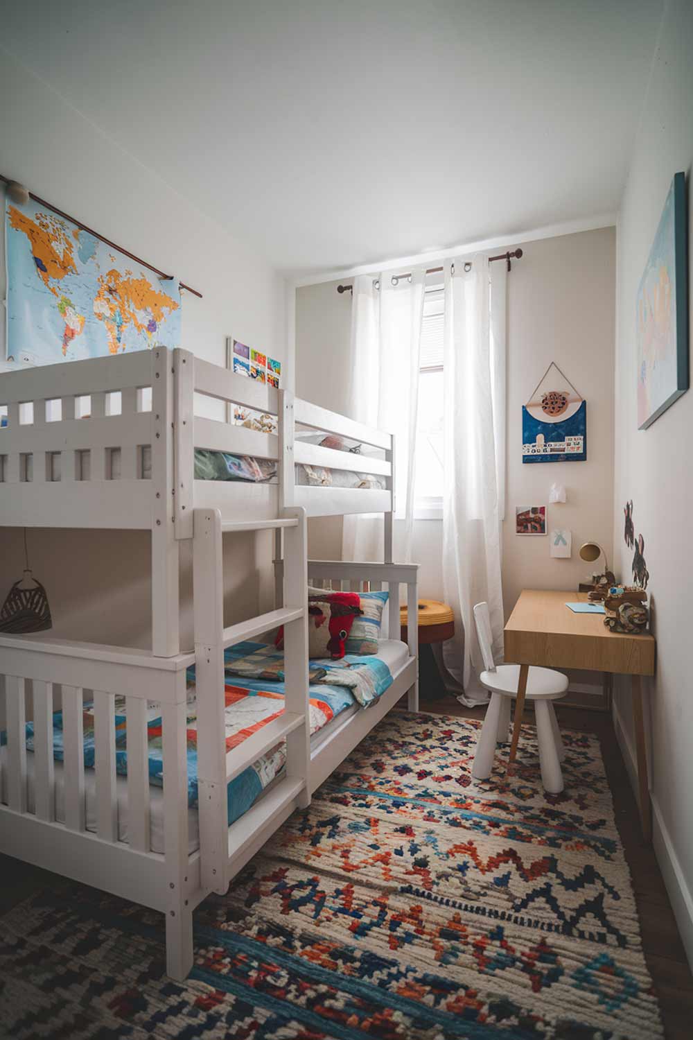 A cozy fun minimalist kids bedroom with a white wooden bunk bed and a small desk. There is a colorful rug beneath the bed. The walls are adorned with a few simple decorations, including a world map and a few paintings. The room has a small window with white curtains.