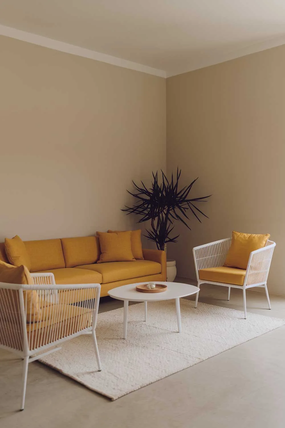 A minimalist living room with a yellow sofa. The room has a white rug and a white coffee table. There are two white chairs with yellow cushions. The walls are beige. There is a potted plant in the corner. The lighting is soft.