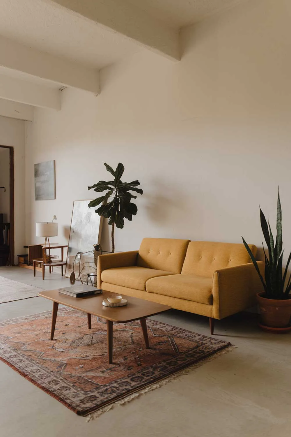 A minimalist living room with a yellow sofa. The room has a white wall, a wooden coffee table, and a potted plant. There's a rug on the floor. The room has a few pieces of decor, including a lamp and a painting. The background contains a doorway.