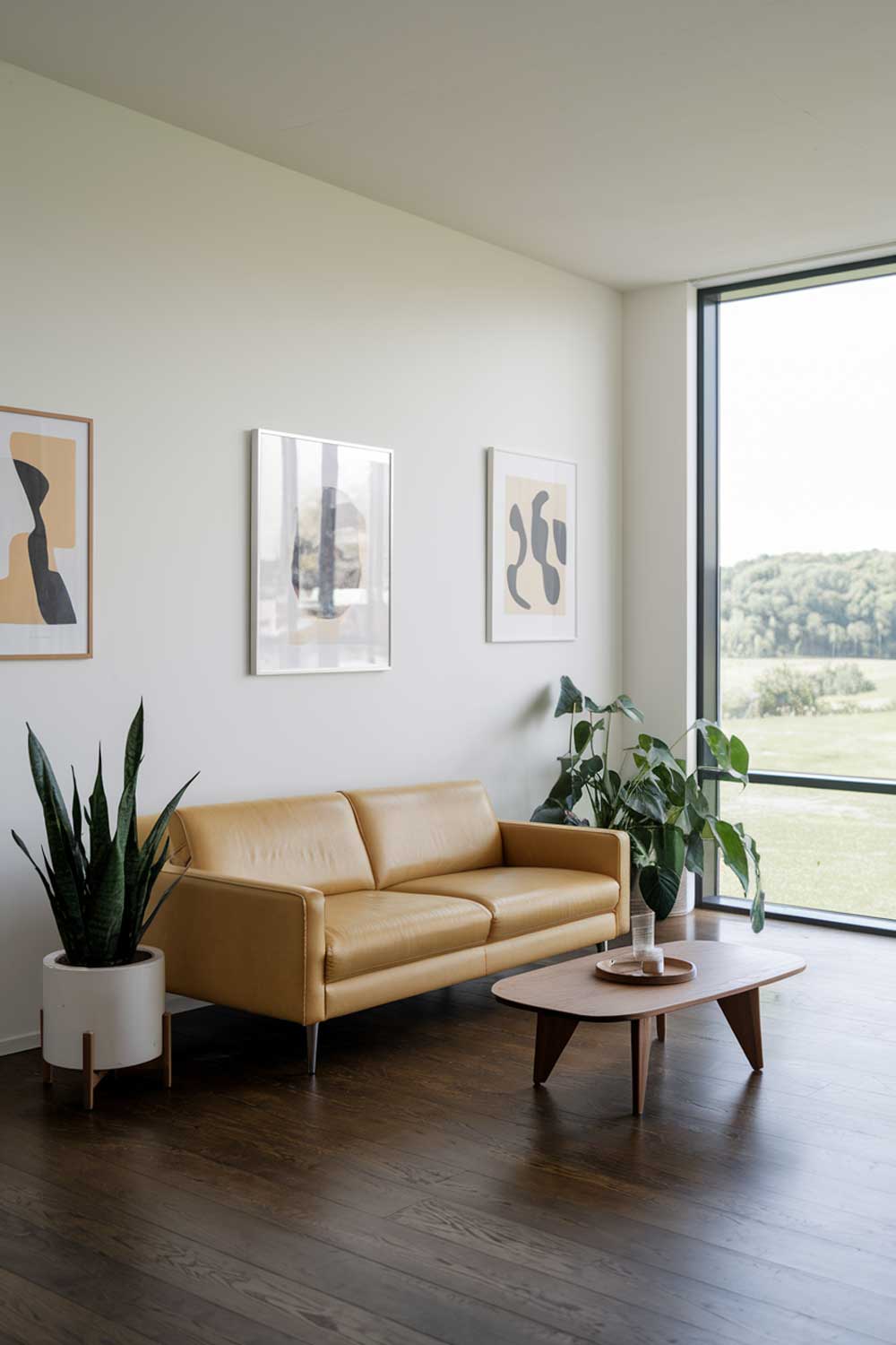 A minimalist living room with a yellow sofa, a wooden coffee table, and a few potted plants. The walls are white, and there are a few pieces of abstract art hanging on the walls. The floor is made of dark wood. The room has a large window with a view of a green landscape.