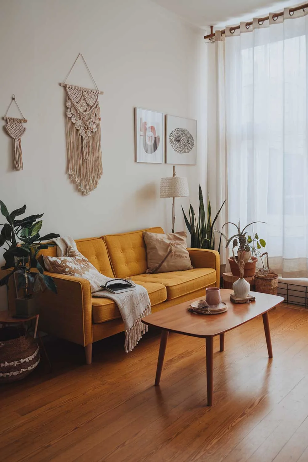 A boho minimalist living room with a yellow sofa, a wooden coffee table, and a few decorative items. There are plants, a throw pillow, and a lamp on the sofa. The walls have a few hanging items, including a macrame wall hanging and a few pieces of art. The room has a wooden floor and a large window with a curtain.