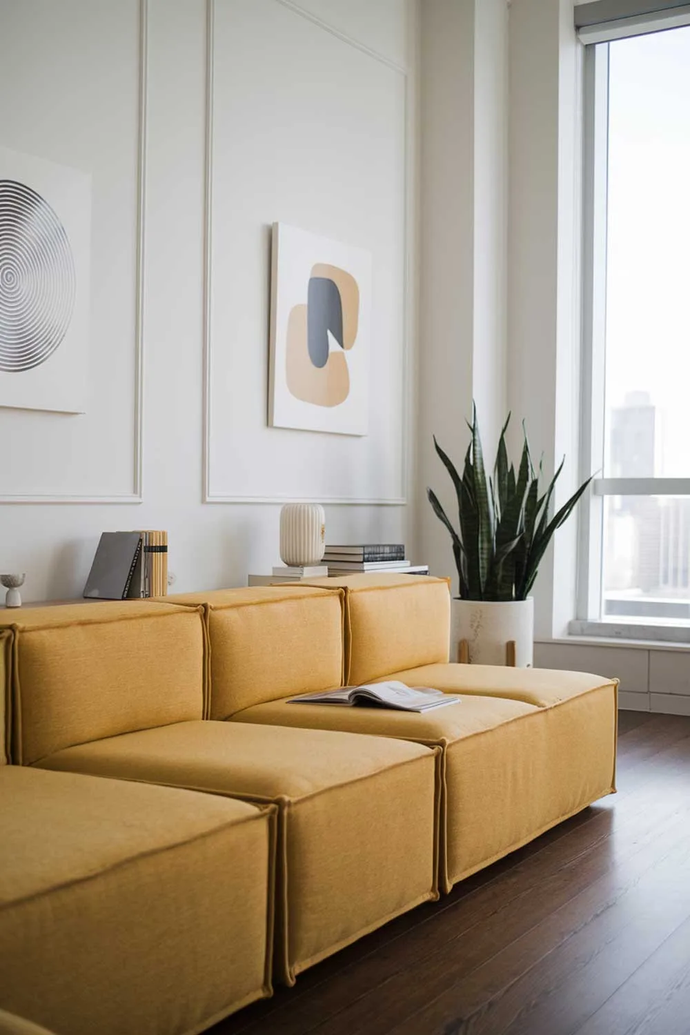 A modern minimalist living room with a yellow modular couch. The room is decorated with a few items, including a small side table, a potted plant, and a few books. The walls are white and have a few pieces of abstract art. There is a large window with a view of a city skyline. The floor is made of dark wood.