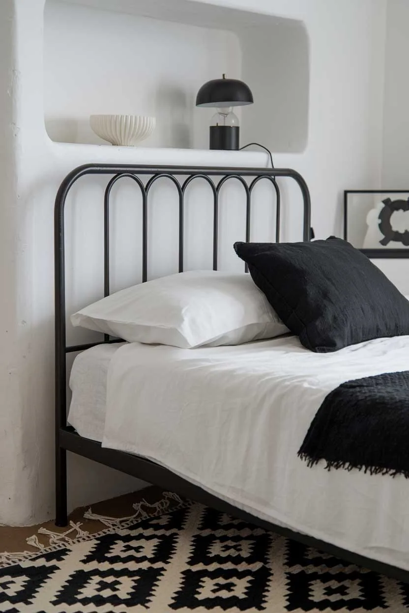 24 Minimalist White and Black Bedroom Ideas 16 A close up of a cozy white and black minimalist bedroom. There's a black metal bed frame with white sheets and a black pillow. There's a white shelf above the bed. On the shelf, there's a black lamp and a white decorative object. The room has a black and white patterned rug. The walls are white.