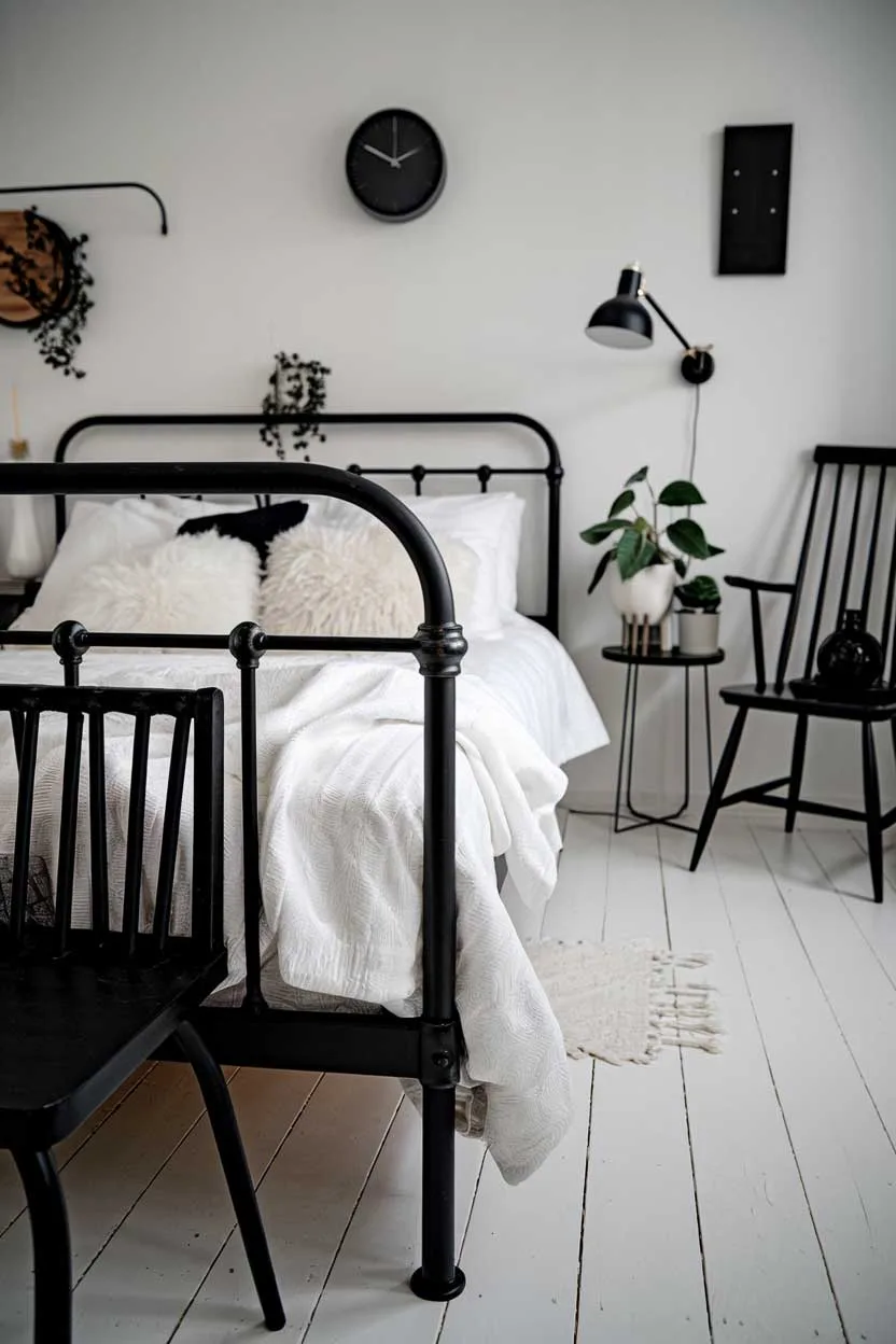 24 Minimalist White and Black Bedroom Ideas 21 A close-up shot of a cozy white and black minimalist bedroom. There's a black metal bed frame with white fluffy pillows and a white blanket. The bed is placed on a white wooden floor. There's a black wooden chair near the bed. The room has a few black decorative items, including a wall clock, a lamp, and a plant stand. The walls are painted white.