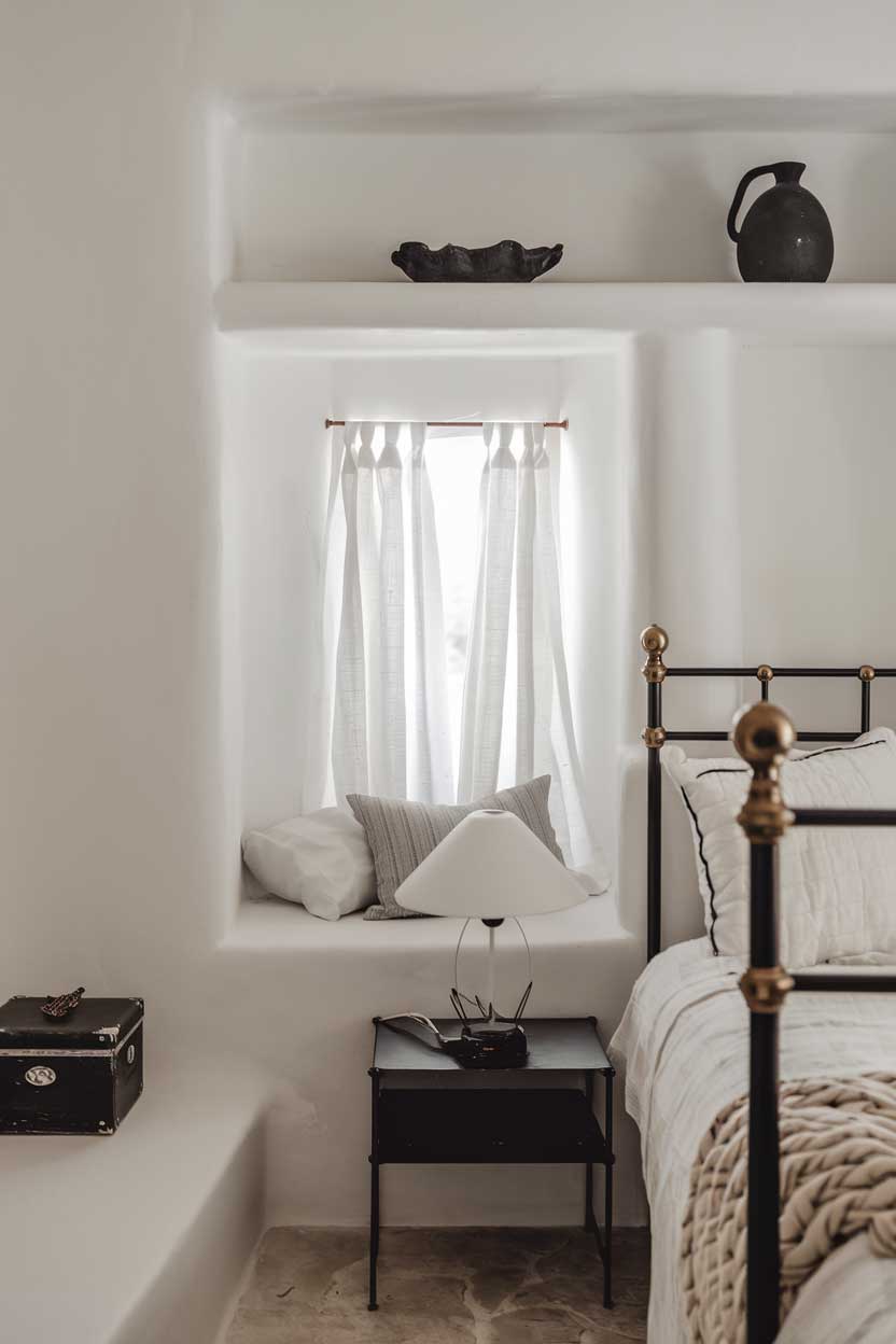 A close-up shot of a minimalist bedroom with a cozy ambiance. The room has a white bed with black frames, a black bedside table, and a white lampshade. Above the bed, there's a white shelf with a few black objects. The walls and floor are white. The room has a small window with white curtains. There's a black box on the floor.