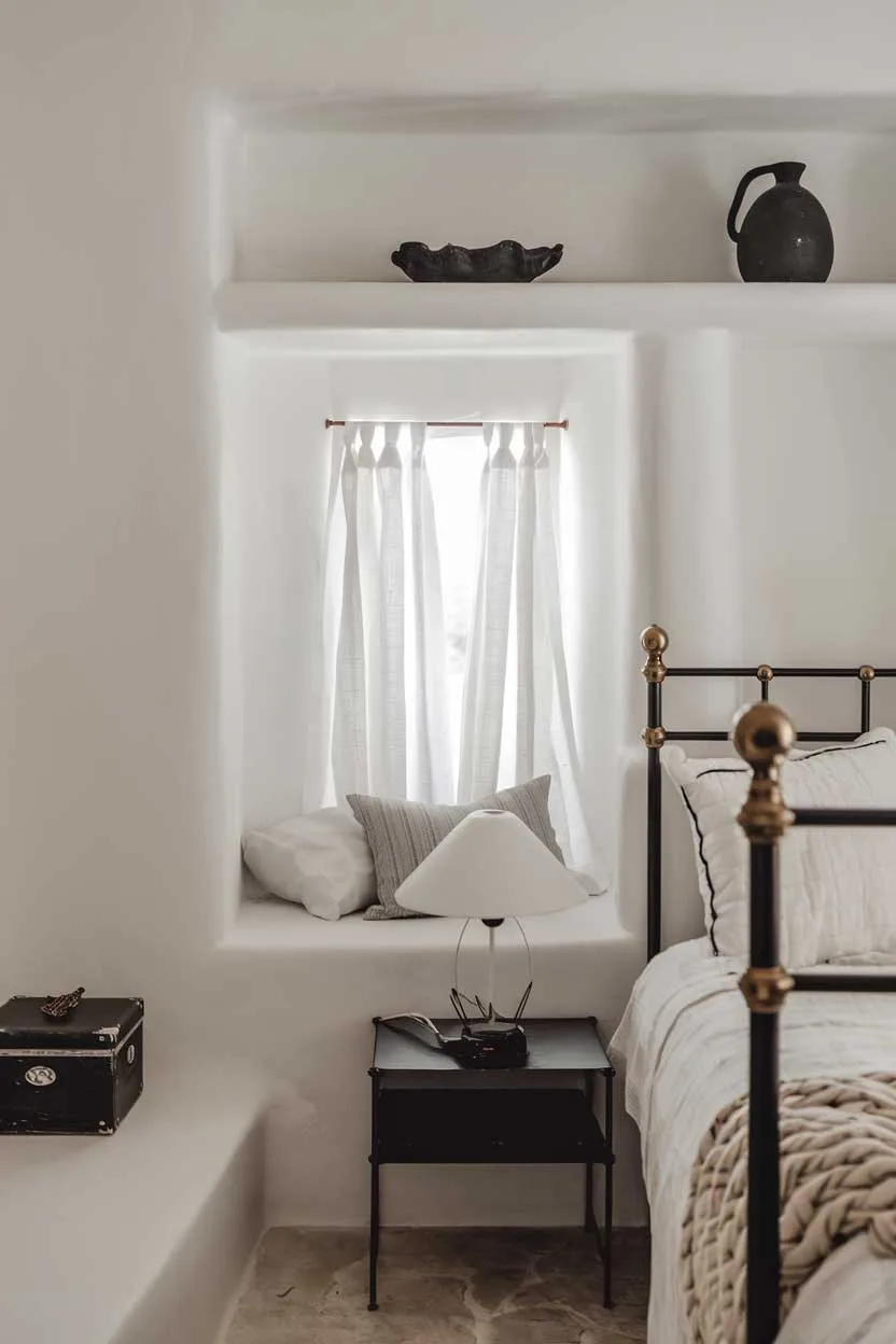 24 Minimalist White and Black Bedroom Ideas 24 A close-up shot of a minimalist bedroom with a cozy ambiance. The room has a white bed with black frames, a black bedside table, and a white lampshade. Above the bed, there's a white shelf with a few black objects. The walls and floor are white. The room has a small window with white curtains. There's a black box on the floor.
