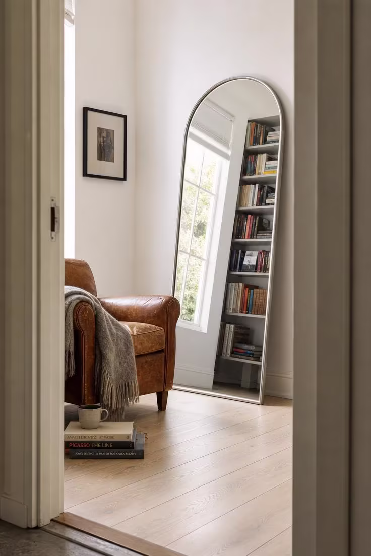 Tall arched floor mirror with brushed steel frame leaning beside a cognac leather reading chair
