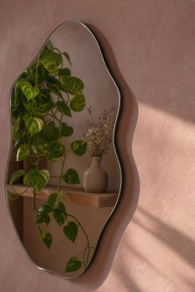 Wavy-edged frameless mirror mounted on a clay-pink limewash wall reflecting a trailing pothos plant