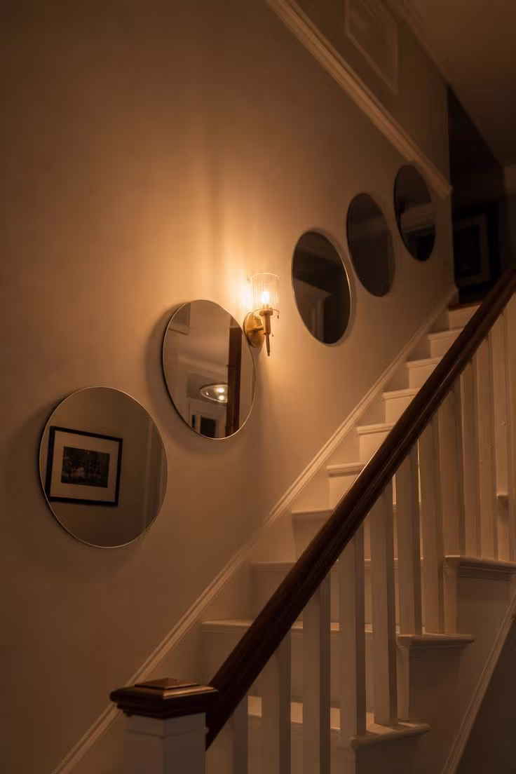 Five identical round frameless mirrors descending along a staircase wall with warm evening sconce light