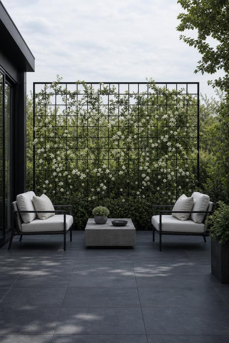 17 Sleek Modern Backyard Landscaping Plans Worth Copying 17 A charcoal porcelain tile terrace with a freestanding black steel trellis covered in star jasmine and two lounge chairs on a cool hazy morning.