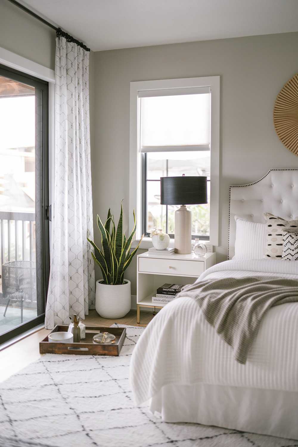 A modern boho bedroom with a stylish decor. The room features a king-size bed with a white headboard. There's a black lampshade on the nightstand. The room has a white rug and a few decorative items, including a plant, a wooden tray, and a few books. The walls are painted in a light gray shade. There's a sliding glass door that leads to a balcony.