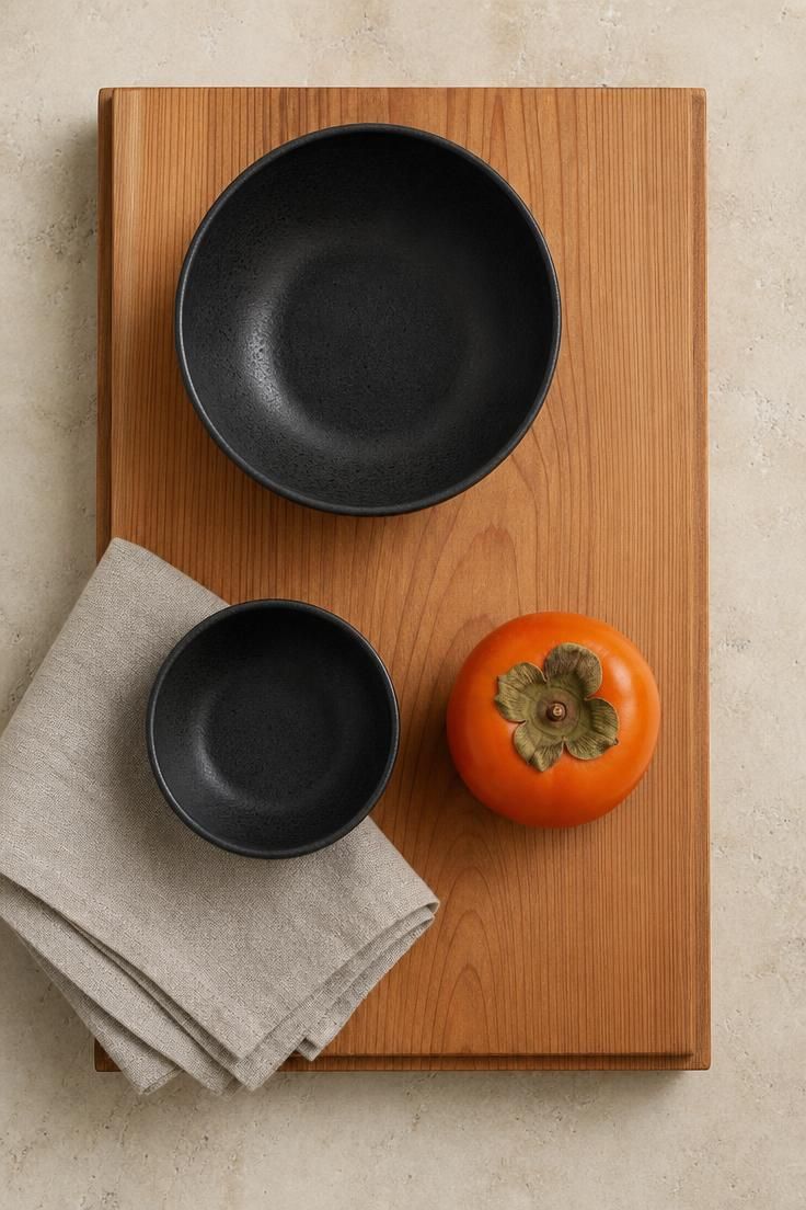 Overhead view of a raw cedar riser holding matte black bowls and a single persimmon in soft diffused light