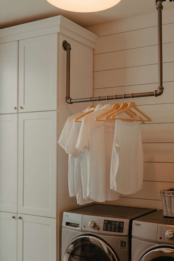 13 Perfect Modern Farmhouse Laundry Room Ideas to Copy 7 Black iron pipe hanging rod mounted between a wall and cabinet with white shirts on wooden hangers in a farmhouse laundry room.