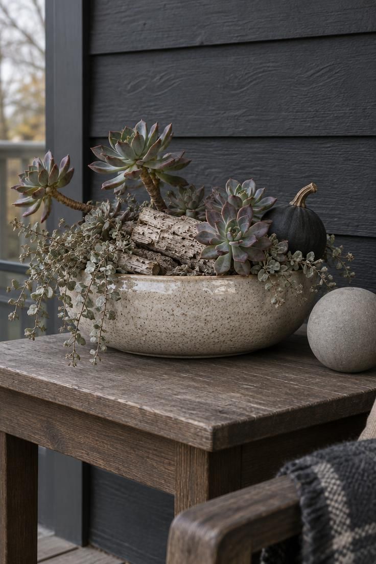 A low speckled ceramic vessel on a charcoal porch table holding woody succulents, pale bark, and one dark gourd, with a concrete sphere beside it under soft overcast light.