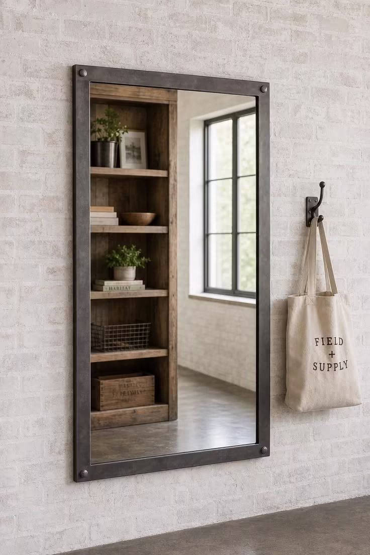Riveted iron-frame mirror on a lime-washed brick wall reflecting reclaimed wood shelving in soft diffused light