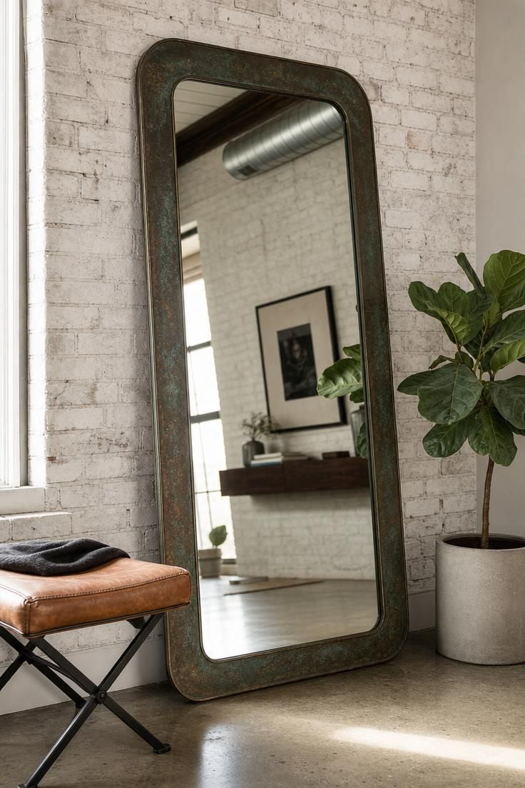 Patinated bronze floor mirror leaning against a whitewashed brick wall next to a fiddle leaf fig in a concrete planter and a saddle leather stool.