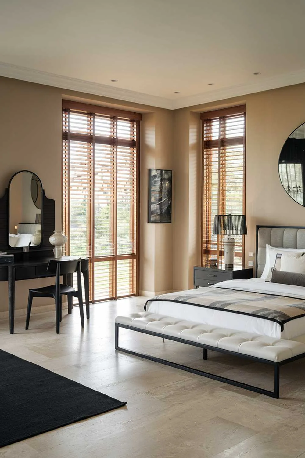 23 Modern Luxury Bedroom Design Ideas You'll Love 4 A modern luxury bedroom design and decor. The room has a white platform bed with a black metal frame and a grey headboard. There is a black wooden dresser with a mirror and a black chair. The room has a large window with wooden blinds. The walls are painted a soft beige colour. The floor is made of beige tiles. There is a black rug near the bed. The room has a few pieces of art on the wall.