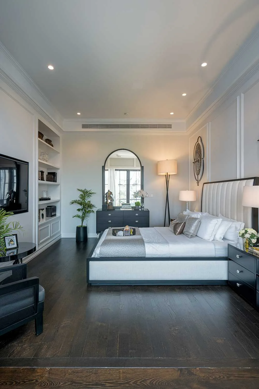 23 Modern Luxury Bedroom Design Ideas You'll Love 8 A modern luxury bedroom design and decor. The bedroom has a king-size bed with a white headboard and a black frame. The bed is positioned against a wall with built-in shelves and a TV. There's a black dresser with a mirror and a black chair next to the bed. The floor is made of dark wood. The walls are painted white. There's a floor lamp near the bed and a table lamp on the dresser. The room has a few decorative items, including a plant, a photo frame, and a small statue. The lighting is soft.