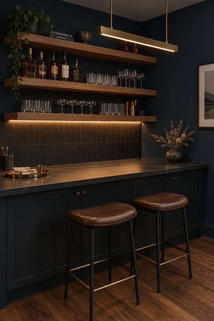 Dark-walled bar corner with floating oak shelves, a honed black stone counter, a brass linear pendant, and two leather bar stools.