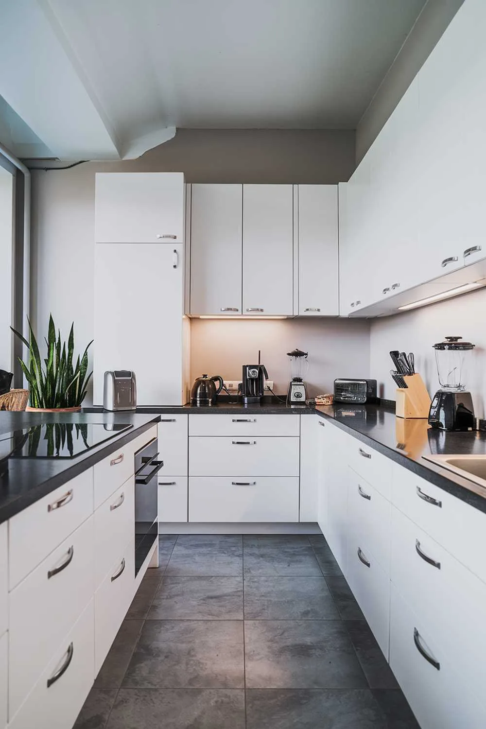 A modern minimalist kitchen with white cabinets, a black countertop, and a few kitchen appliances. There is a black kettle, a toaster, and a blender on the countertop. There are also a few cooking utensils. The floor is made of gray tiles, and the walls are painted in a light gray hue. A potted plant is placed near the window. The lighting is bright.