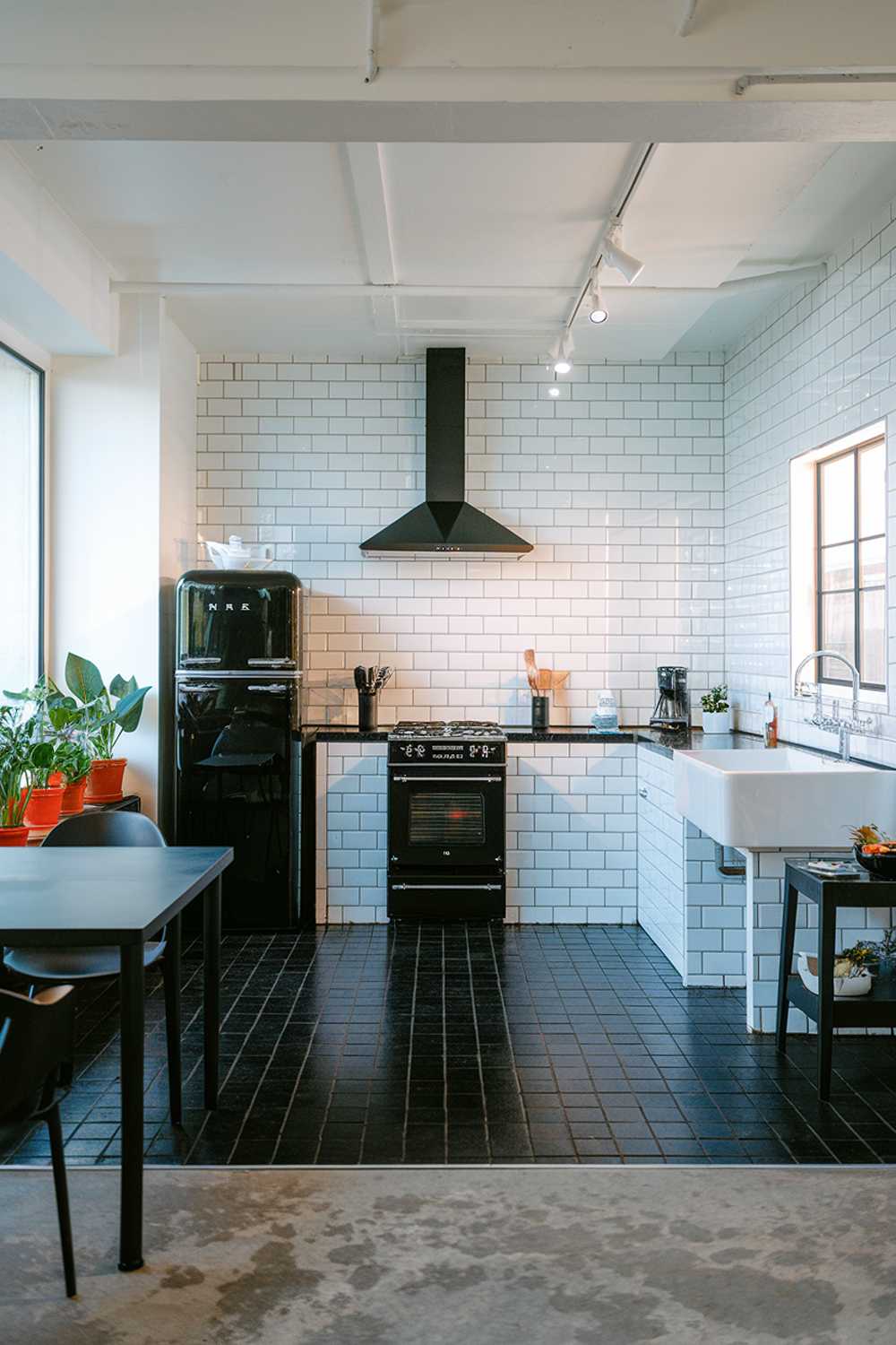 A modern minimalist kitchen design. The kitchen has a white subway tile backsplash, a white countertop, and a white sink. There is a black refrigerator, a black stove, and a black table. The floor is made of black tiles. There are some potted plants near the window. The lighting is bright.