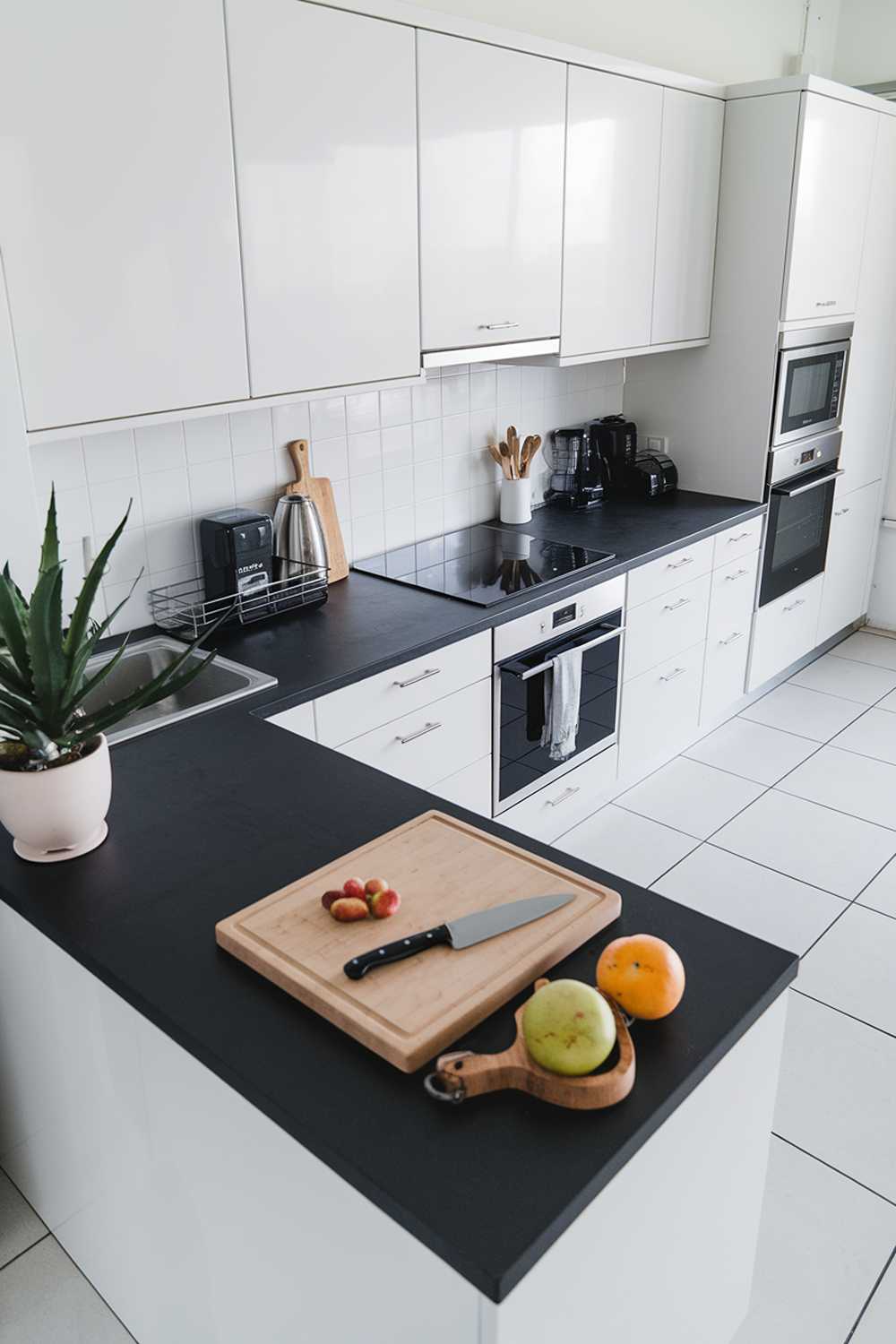 A modern minimalist kitchen design with a black countertop, white cabinets, and a few stainless steel appliances. There's a wooden cutting board with a knife and some fruits on the countertop. A potted plant is placed beside the sink. The floor is covered with white tiles.