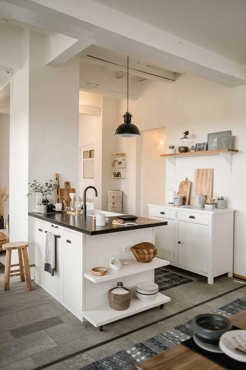 A cozy modern minimalist kitchen design. There's a white kitchen island with a black countertop, a sink, and a few items. There's a white cabinet with a few items on top. There's a black pendant light hanging over the kitchen island. The floor is made of gray tiles. The walls are painted white. There's a wooden stool near the kitchen island. The space is well-lit.