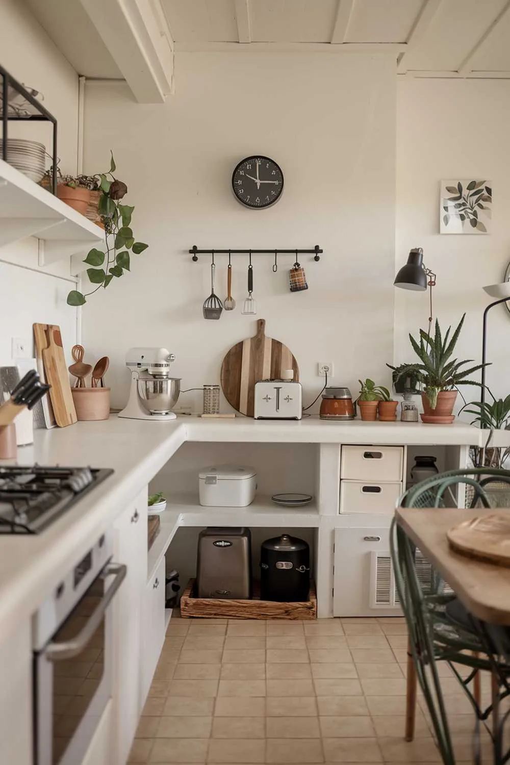  A modern minimalist kitchen design. The kitchen has a white countertop with a few appliances, including a mixer and a toaster. There's a wooden cutting board and a few utensils near the mixer. On the wall above the countertop, there's a black clock and a few hooks. The floor is covered with beige tiles. The room has a few potted plants and a lamp.