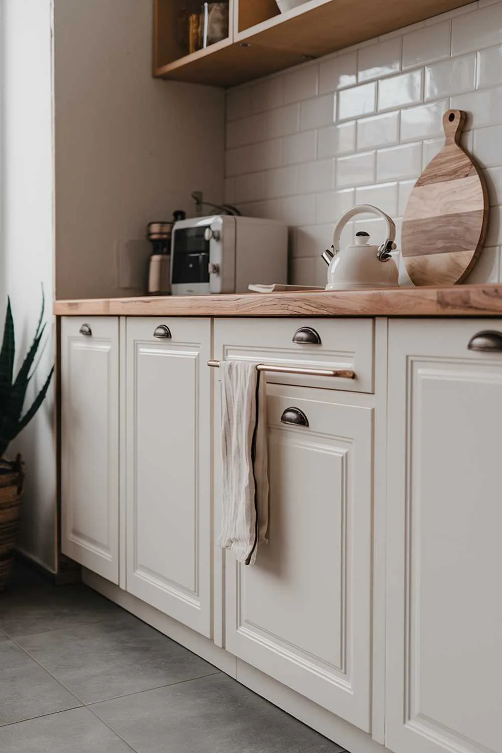 A modern minimalist kitchen design. The kitchen has a white cabinet and a wooden countertop. There is a white kettle and a wooden cutting board on the countertop. There is a potted plant near the cabinet. The floor is covered with gray tiles.