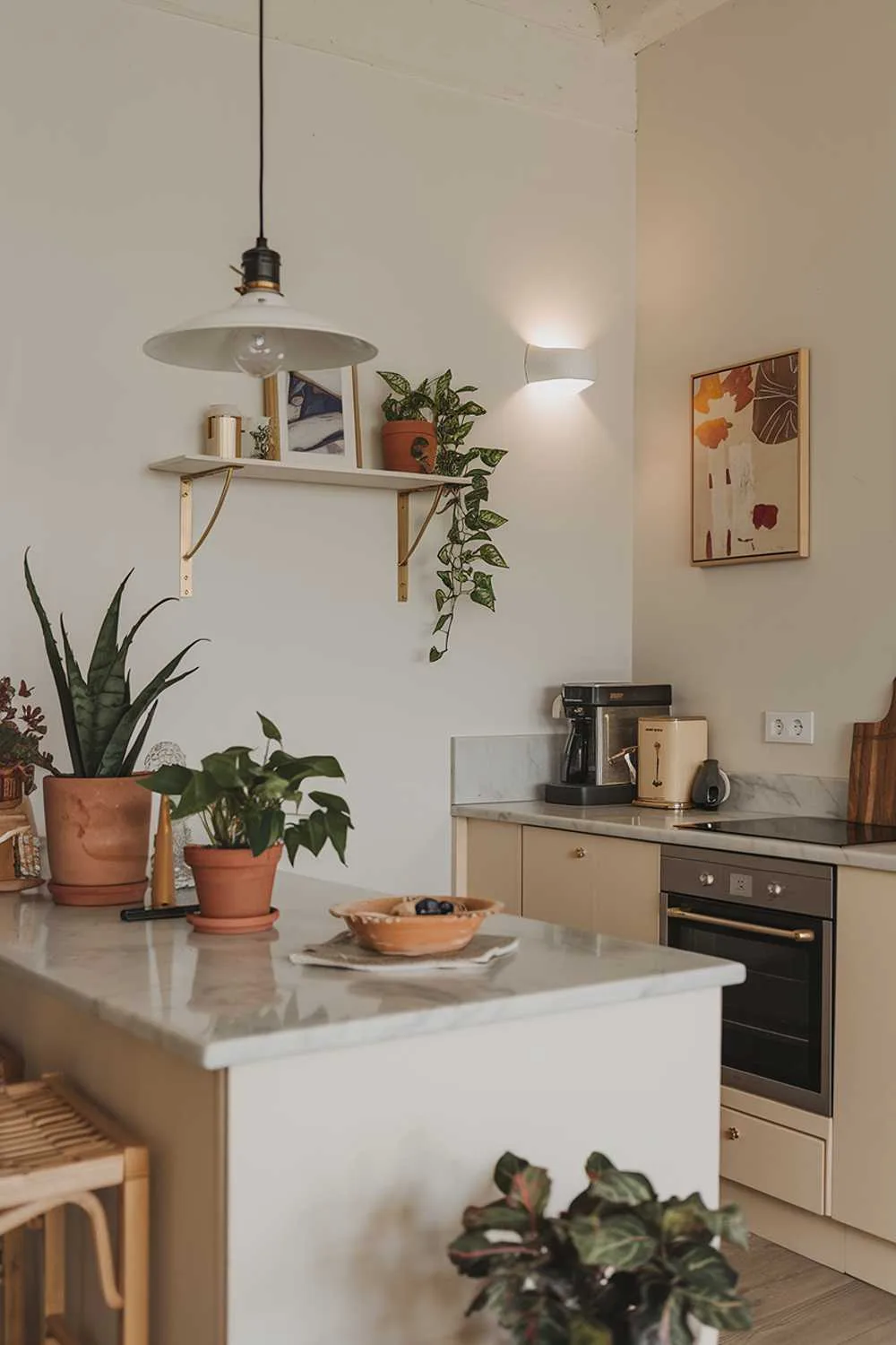 A cozy modern minimalist kitchen design with a few key elements. There's a clean white island with a marble countertop, a few potted plants, a few decorative items, and a pendant light. There's a wall oven and a cooktop. On the wall, there's a floating shelf with a few items, a piece of art, and a sconce light. The floor is a light wood.