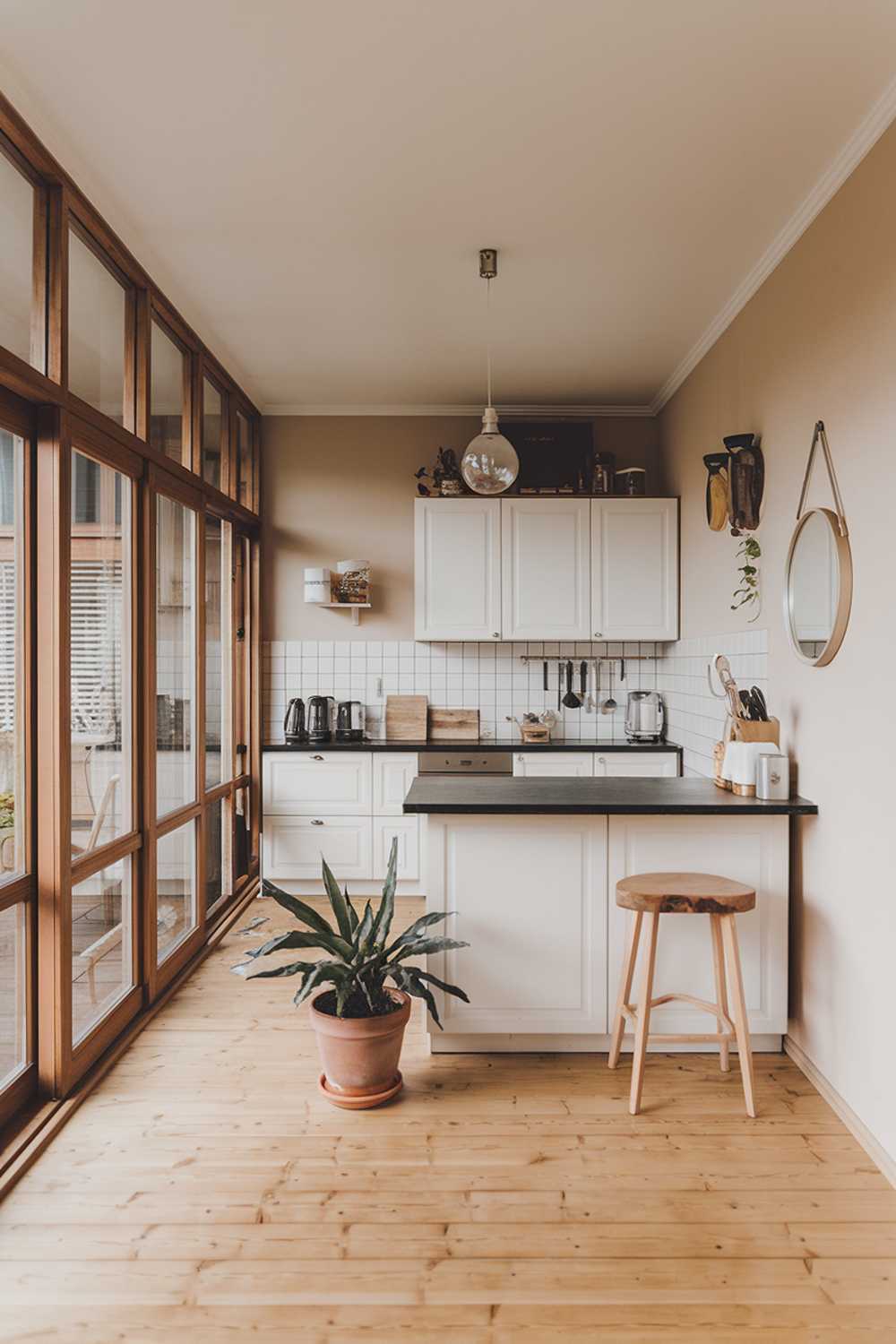 A modern minimalist kitchen design. The kitchen has a wooden floor and is furnished with a white kitchen island, white cabinets, and a black countertop. There is a wooden stool beside the island. A potted plant is placed on the floor. A few utensils are placed on the black countertop. The room has a large window with wooden frames. The walls are painted beige. A few personal items, like a mirror and a decorative object, are placed on the beige wall.