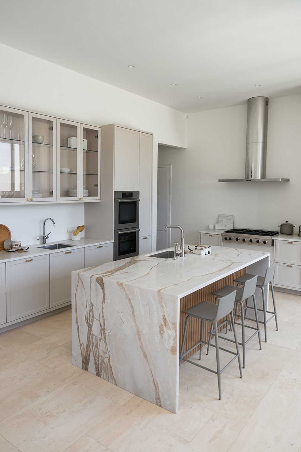 A modern minimalist kitchen design. There's a large island in the middle with a marble countertop and a few bar stools. On the left, there's a white cabinet with glass doors, housing some kitchenware. Next to it is a sink with a faucet. On the right, there's a stovetop with four burners and a chimney above it. The floor is covered with beige tiles. The walls are painted white.