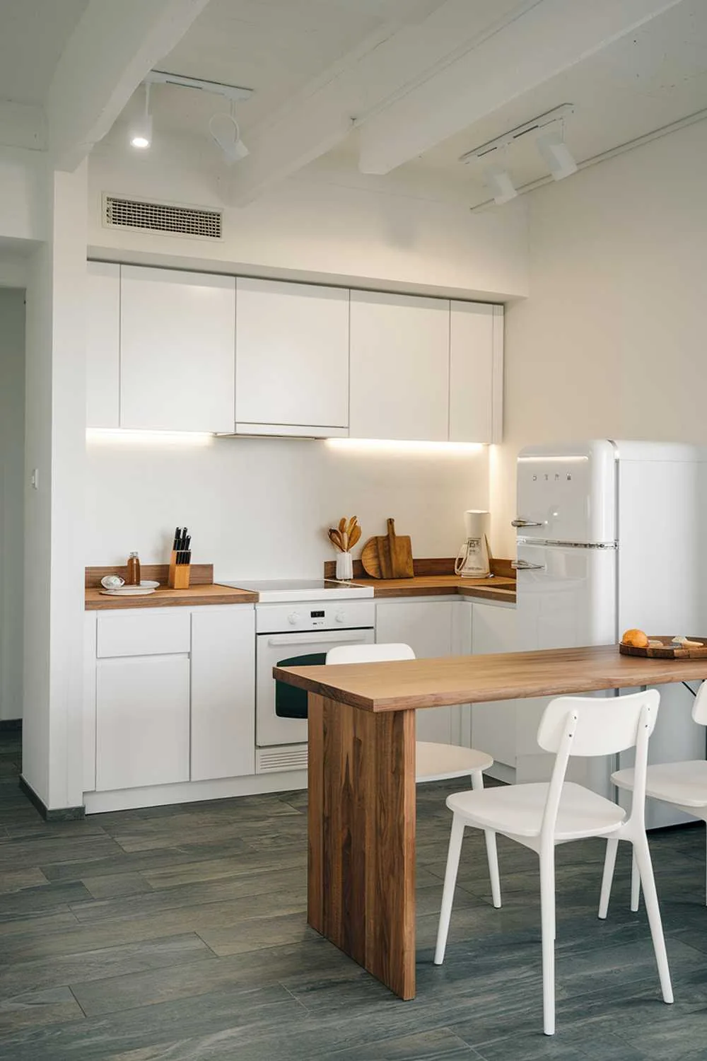  A modern minimalist kitchen design. The kitchen has a white color scheme with wooden elements. There is a white cabinet with a wooden countertop, a white oven, and a white refrigerator. There is a wooden table with two white chairs in front of the refrigerator. The floor is made of gray tiles. The lighting is bright.