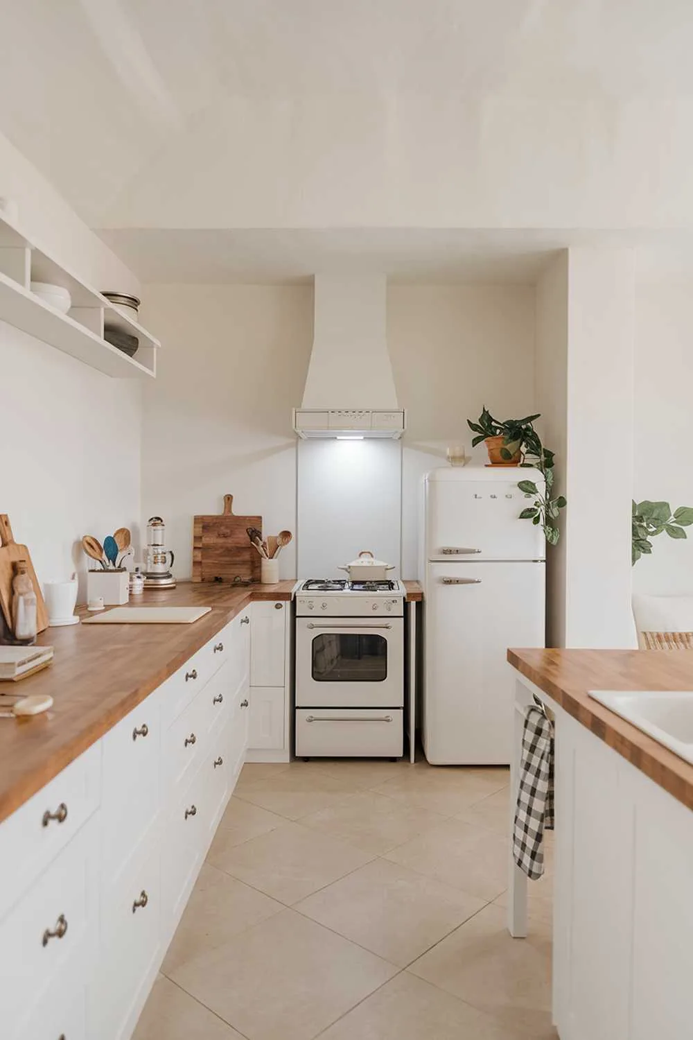 A modern minimalist cozy kitchen design. The kitchen has a white cabinet and a wooden countertop. There's a white stove, a white refrigerator, and a wooden cutting board. There are a few utensils and a pot on the counter. The floor is covered with beige tiles. The walls are painted white. There's a potted plant near the refrigerator. The lighting is bright.