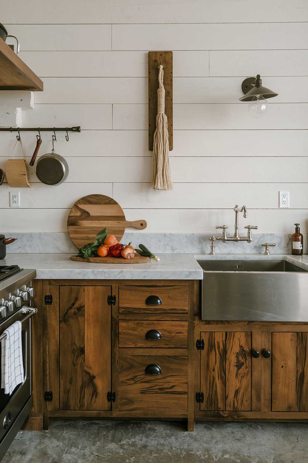 A modern rustic kitchen with wooden cabinets, a marble countertop, and a stainless steel sink. There is a wooden cutting board with vegetables on the countertop. Hanging above the sink is a decorative item made of wood and rope. The floor is made of concrete. The walls have a few items hung on them, including a pot, a pan, and a light fixture.